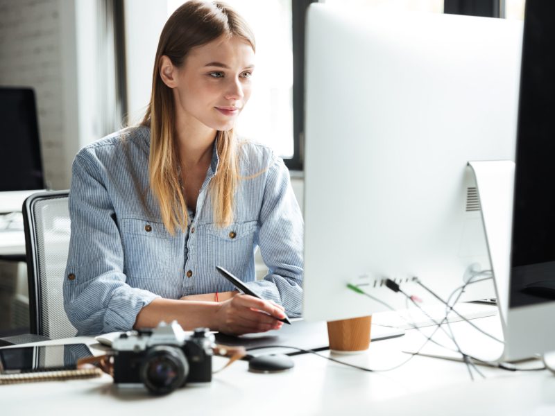 Photo of cheerful young woman work in office using computer and graphic tablet. Looking aside.